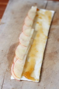 a strip of pastry on a cutting board with a line of apples on one side and jam covering the pastry